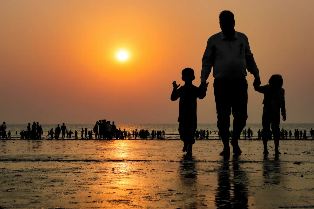 : Sunset silhouette of a father walking on the wet beach sand holding hands with his two children.